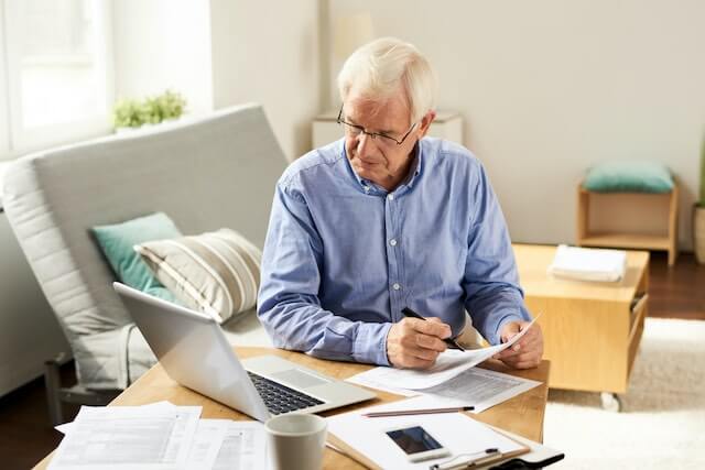 Senior aged man sitting at a desk at home with paperwork spread in front of him as he looks at a laptop and fills out a paper form