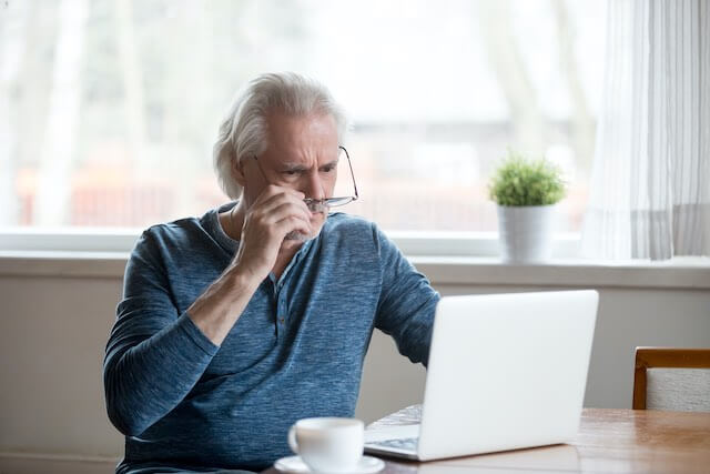 Senior aged man staring at a laptop while sitting at his desk with a shocked, frustrated look on his face as he pulls his glasses down just beneath his eyes while staring at the computer screen in disbelief