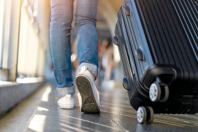Close up of the legs and feet of a person wearing blue jeans pulling a suitcase on wheels through an airport