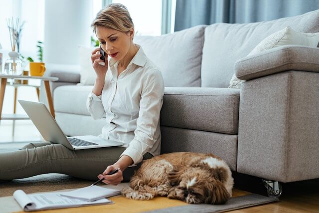 Woman pictured working at home sitting on the floor in front of a couch with a laptop while she talks on the phone and looks at paperwork; a dog is sleeping beside her on the floor