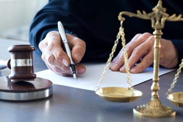 Close up of a judge's hands signing a document while he sits at his desk; justice scales and a wooden gavel are seen in the foreground