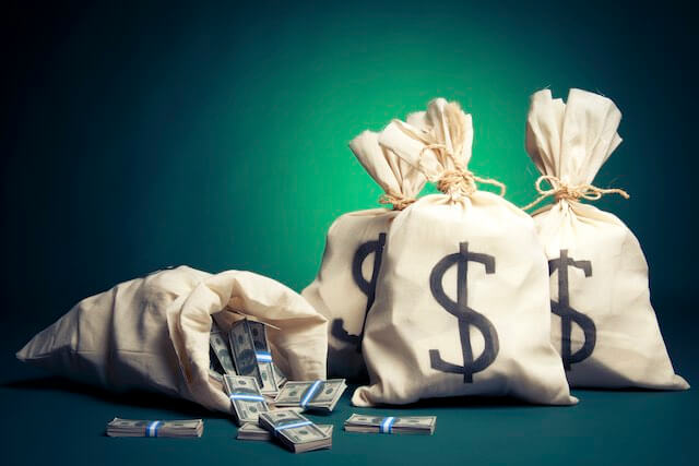 Bags of cash against a dark green background; one is lying open on its side and has some cash lying in front of it