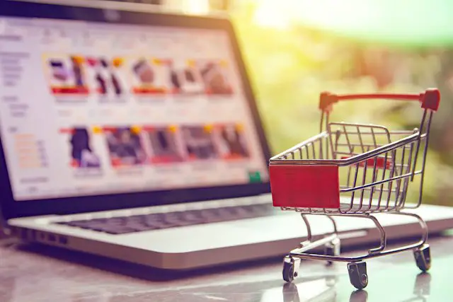 Miniature shopping cart pictured on a desk in front of a laptop depicting online shopping/e-commerce