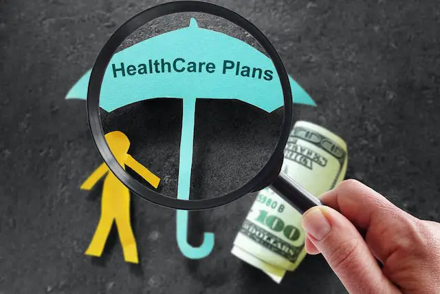 Close up of a person's hand holding a magnifying glass over a paper umbrella on a dark surface that reads 'healthcare plans' pictured next to a cut out of a paper person and a rolled up $100 bill