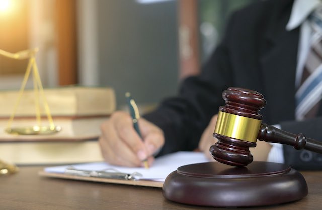 Judge/lawyer sitting at a desk writing with a gavel pictured in the foreground and law books in the background