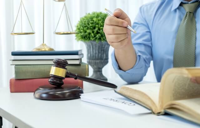 Close up of a lawyer at a desk looking through an open book with a document in front of him labeled 'contract'; a wooden judge's gavel and justice scale on a stack of law books is pictured off to the side