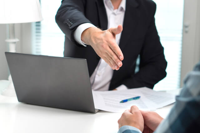 Businessman extending his hand offering a handshake to a man sitting across the desk; there is a laptop open and paperwork on the desk in between