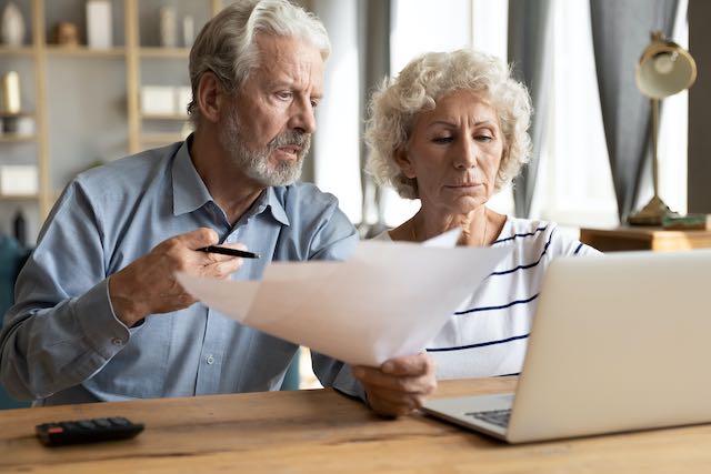 Senior aged couple sitting at a computer with paperwork appearing hassled/stressed as they study taxes/finances