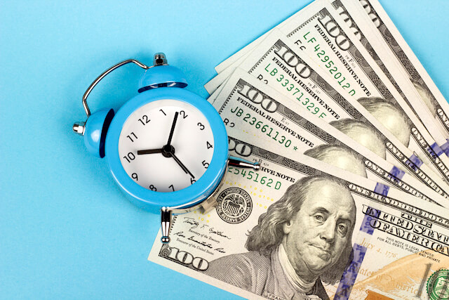 Old style blue alarm clock next to a spread of $100 cash bills on a blue background