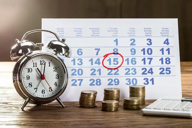 Old style alarm clock, stacks of coins and a calculator pictured in front of a desktop paper calendar with the date 15 circled on it