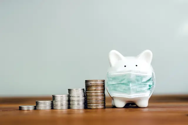 Piggy bank on a wooden surface wearing a face mask next to vertical stacks of coins increasing in size from left to right depicting investing/saving money/retirement/emergency fund