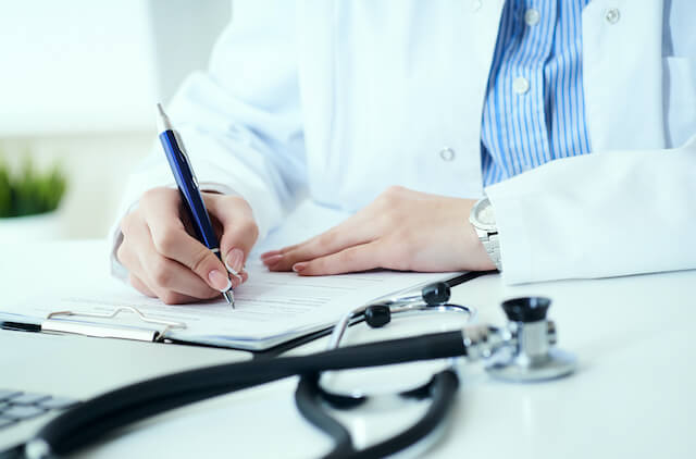 Close up of a doctor's hands filling out paperwork while sitting at a desk; a stethoscope is seen lying in the foreground