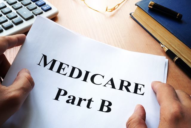 Close up of a person's hands holding a sheet of paper on a desk that reads 'Medicare Part B'; a calculator, book and reading glasses are seen lying off to the side