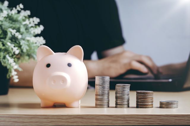 Pink piggy bank next to vertical stacks of coins on a desk growing in size from right to left; a man is seen working on a laptop in the background