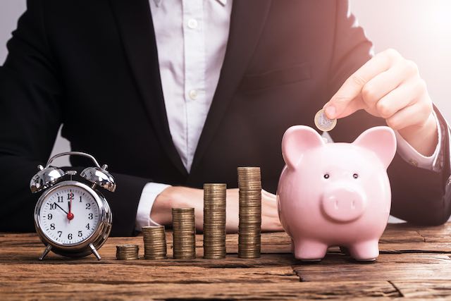 Businessman's hand putting money into a pink piggy bank pictured on the end of five stacks of coins growing in size from left to right next to an old style alarm clock - savings growth over time