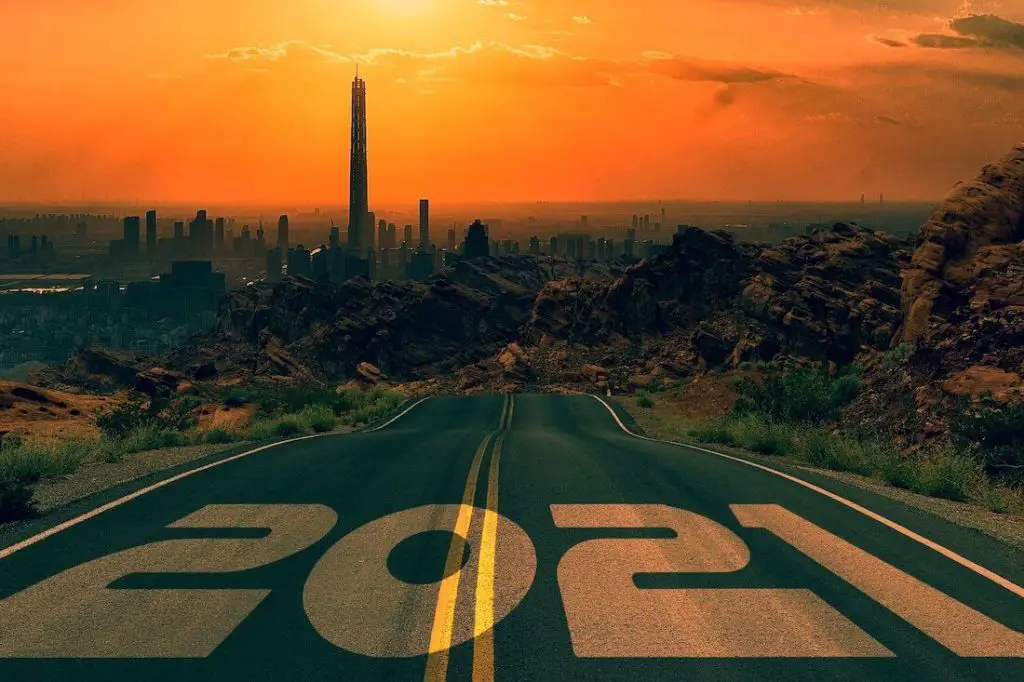 Two lane road leading towards a city skyline in the background against an orange sunset with the numbers '2021' illustrated over the top of the highway