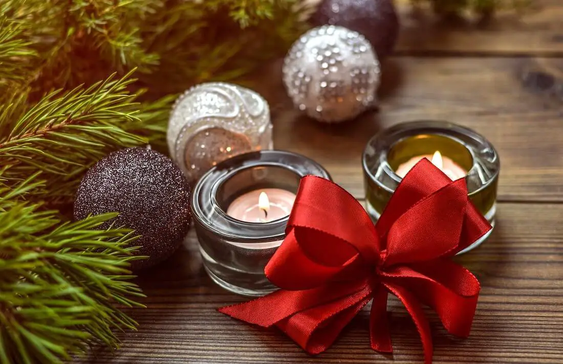 Two candles adorned with a red Christmas bow next to pine branches and two Christmas ornaments on a wooden surface