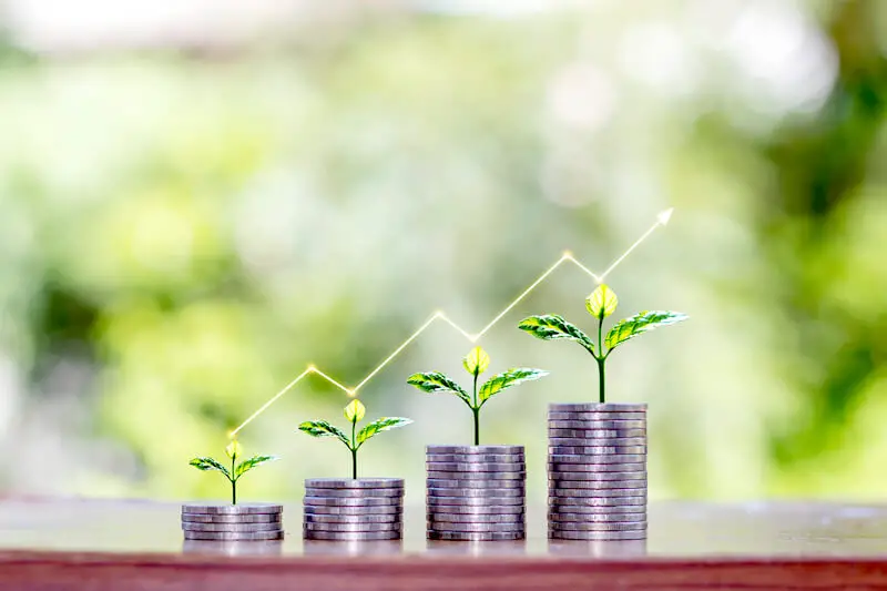 Four vertical stacks of coins growing in size from left of right with plants growing out of the top of each with a trend line pictured above moving in an upward direction