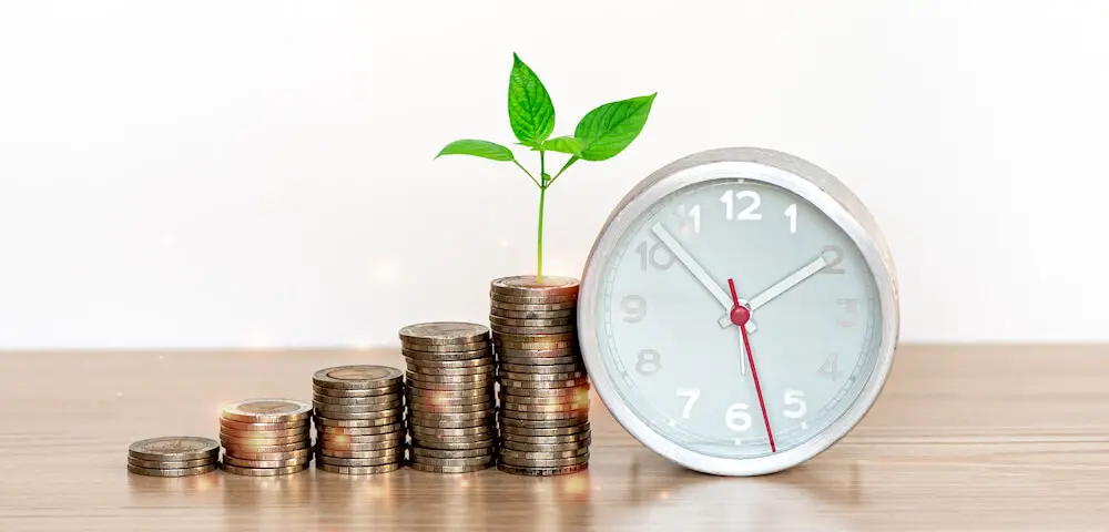 Vertical stacks of coins growing in size from left to right on a wooden surface with a plant growing out of the top of the tallest stack; a clock is pictured next to the coins