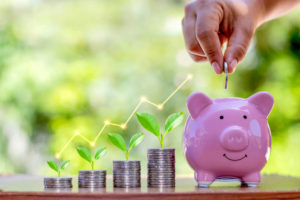 Vertical stacks of coins growing in size from left to right with an upward trend line arrow above them and a pink piggy bank on the right with a close up of a person's hand putting a coin into it