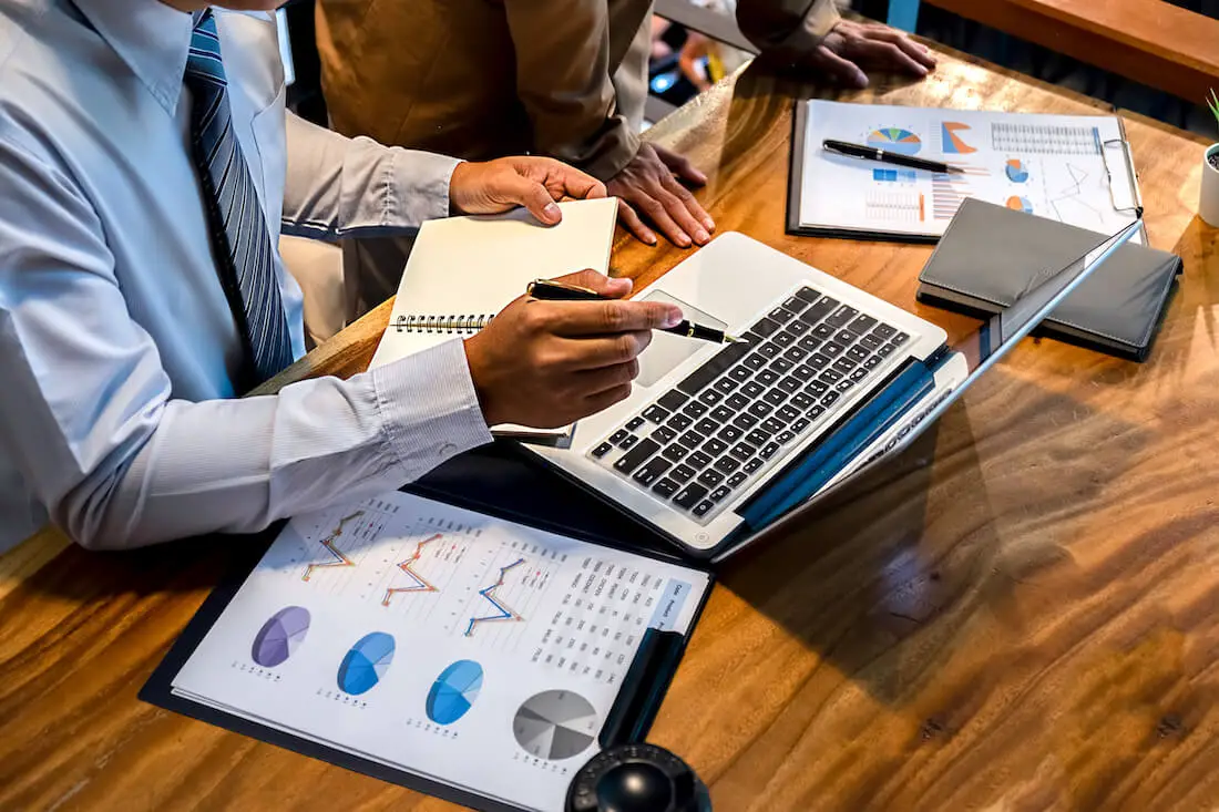 Two businessmen reviewing spreadsheets and reports on a laptop at a desk