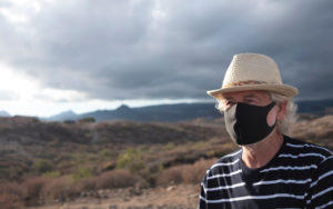 Senior aged man wearing a black facemark outdoors with hills and cloudy skies pictured behind him