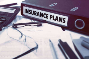 Binder labeled 'insurance plans' next to glasses, pens and paperwork