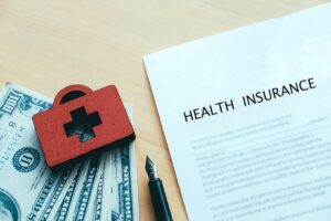 Paper form labeled 'health insurance' next to cash, a pen, and a red medical first aid kit symbol