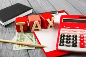 Block letters spelling 'tax' on a desk next to cash, a notepad, pencil and calculator