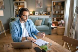 Man engaged in telework at home while sitting at a desk in front of a laptop and taking notes on a notepad