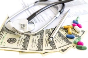 Stethoscope sitting on top of health insurance paperwork with a spread of $100 bills, a pen and some pills