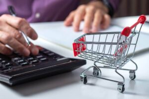 Close up of a person performing calculations on a calculator and writing notes on a notepad with a small grocery shopping cart on the desk in the foreground