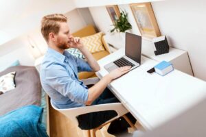 Man teleworking sitting at a desk in the corner of a bedroom working on a laptop while talking on a phone