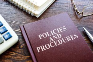 Book titled 'policies and procedures' lying on a desk next to a calculator, pen and reading glasses