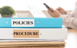 Two binders labeled 'policies' and 'procedure', respectivey, on a desk with a person working on a tablet in the background