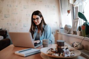 Woman working on a laptop at her kitchen table. A window is behind her and a coffee cup and notepad with pen are on the table next to the computer