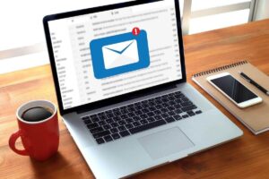 An email icon with a new message notification is pictured on a laptop screen; the computer is on a desk next to a coffee cup, notepad, pen and smartphone
