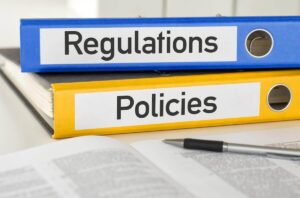 Binders labeled 'regulations' and 'policies', respectively, on a desk with a document and pen in front of them