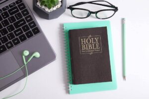 Copy of the Bible on a desk next to reading glasses, a laptop, and earbuds