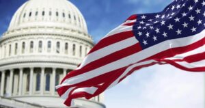 USA flag flying in the wind with the U.S. Capitol building pictured behind it against a blue sky