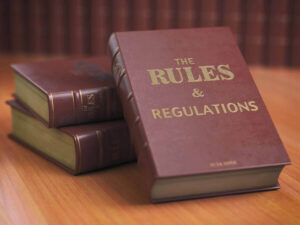 Books on a desk with one facing up that says "The Rules & Regulations" on the cover